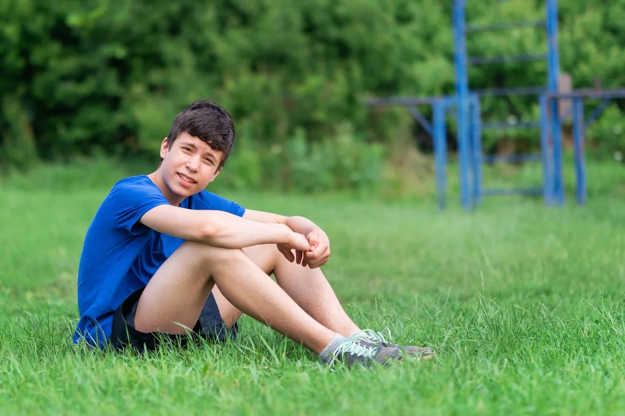 A young boy sitting on the grass in a park, wearing a blue t-shirt and shorts, with a playground in the background.