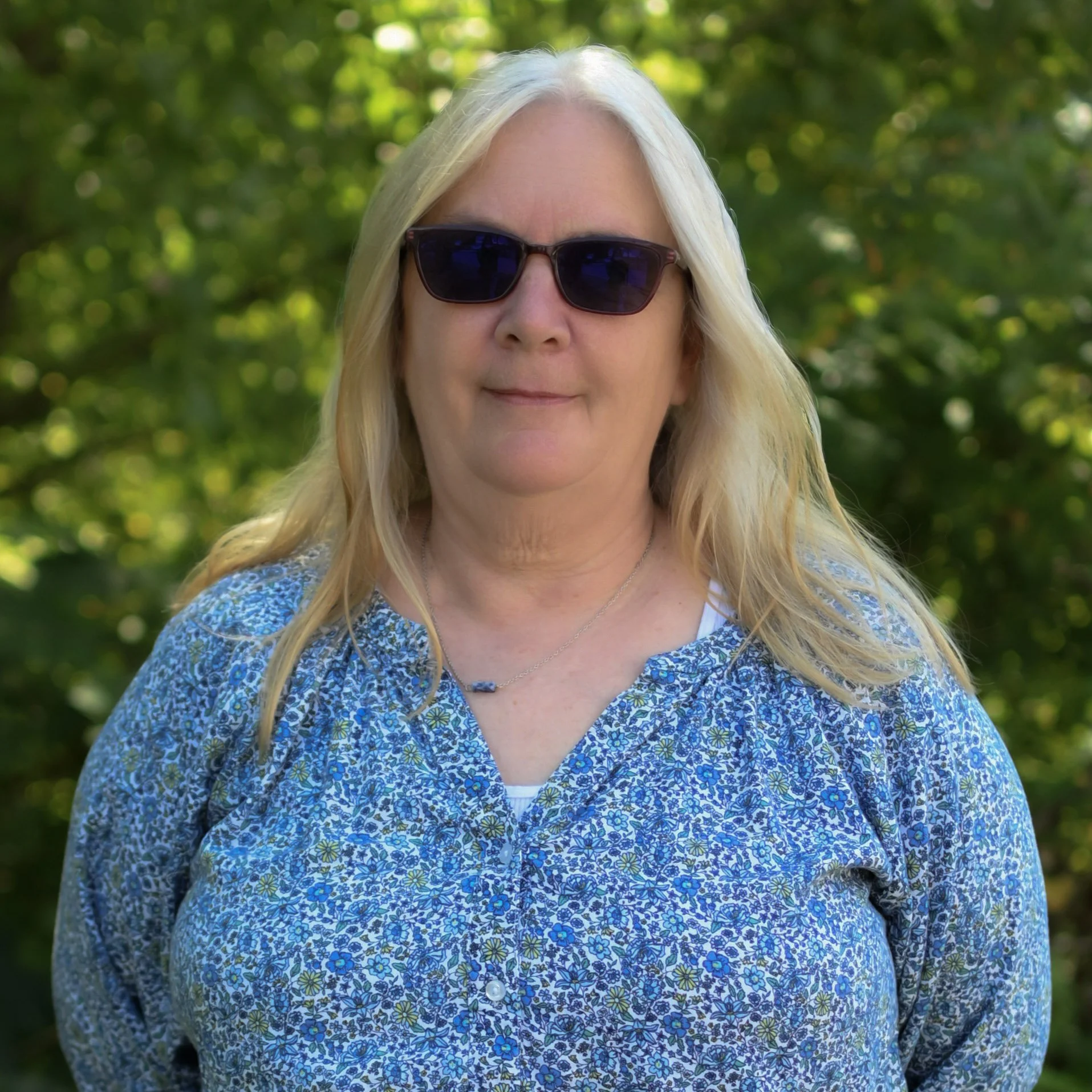 A woman with blonde hair wearing dark sunglasses and a blue floral blouse, standing outdoors with green trees in the background.