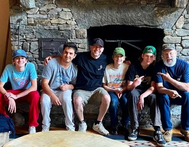 Six people sitting on a wooden bench in front of a stone fireplace, smiling and posing for the photo.