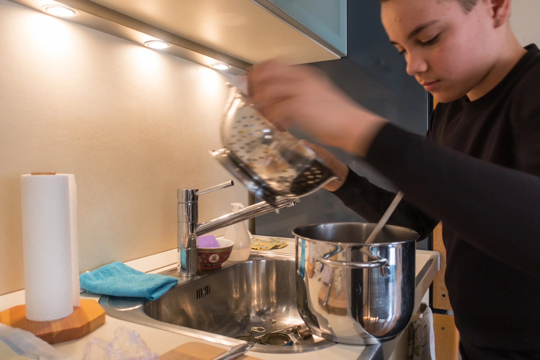 A young person pouring contents into a large stainless steel pot on a kitchen countertop, with a paper towel roll, a blue cloth, and kitchen items around the sink.