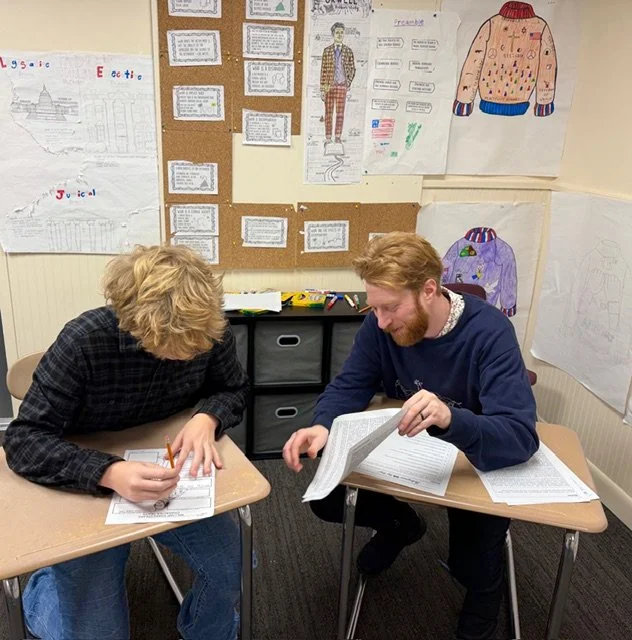 Two young men working together at desks in a classroom, reviewing papers. The classroom has posters, drawings, and a corkboard wall with various artwork and notes.
