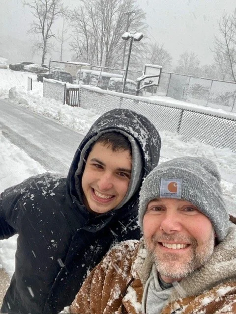 A smiling young man and an older man outdoors in a snowy environment, both wearing winter hats and jackets, with snow on their clothing and a snow-covered tennis court and fence in the background.