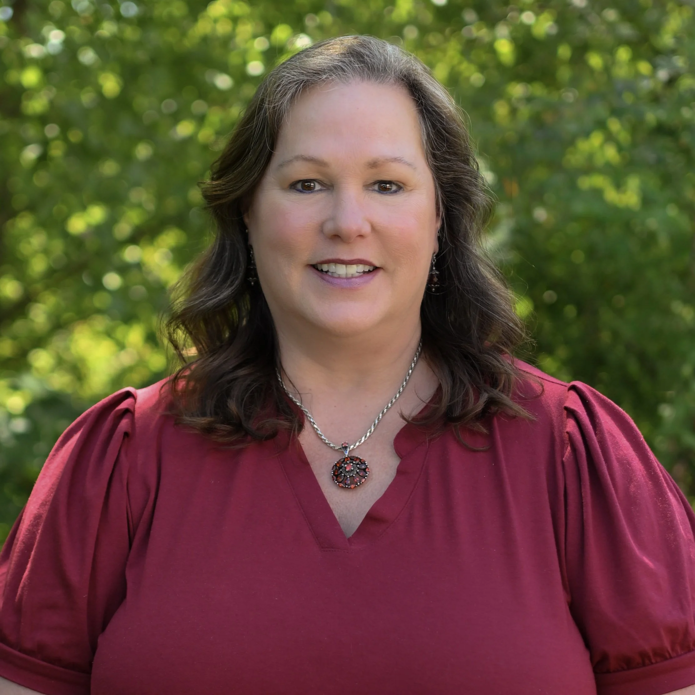 Portrait of a woman with brown hair, wearing a red blouse, silver jewelry, and earrings, standing outdoors with green foliage in the background.