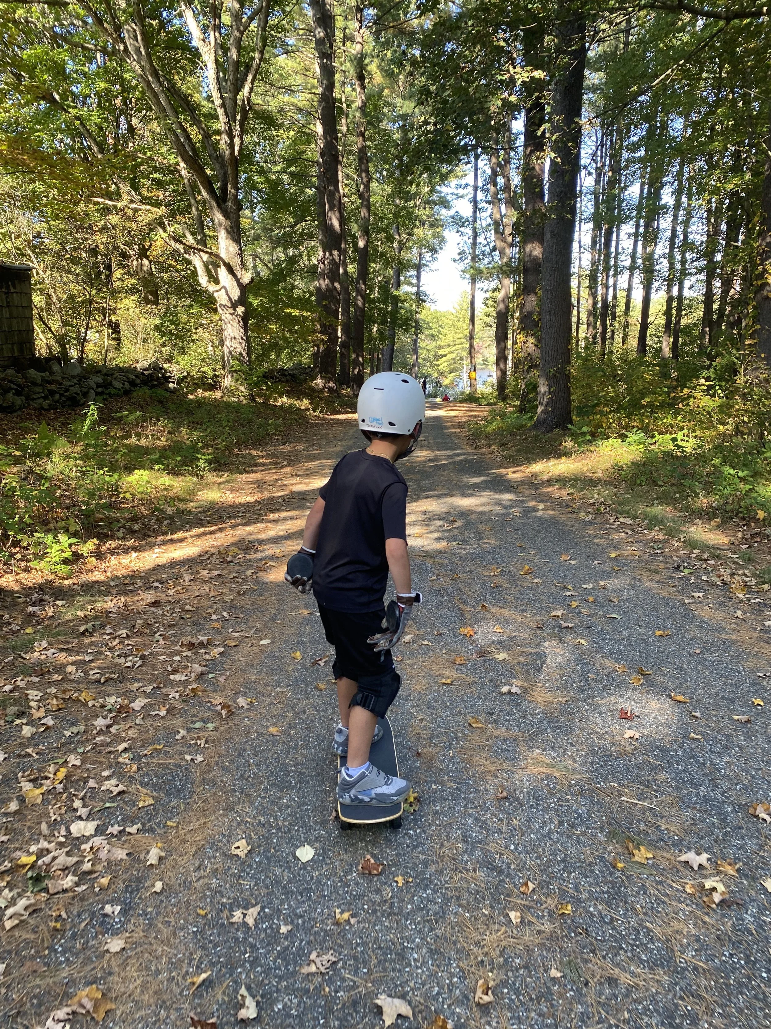 A young boy skateboarding on a gravel trail surrounded by trees and foliage in a wooded area.