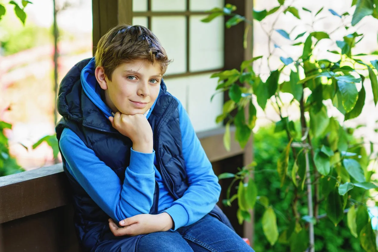 A young boy with brown hair and blue eyes sitting on a wooden railing, resting his chin on his hand, wearing a blue hoodie and a dark blue vest, outdoors near greenery and a window.