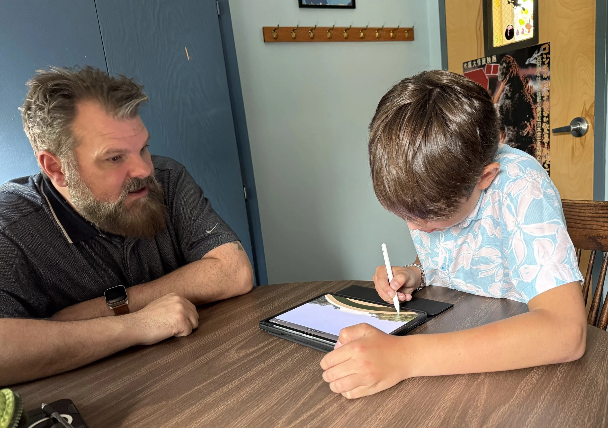 A boy using a stylus on a tablet with a landscape image, while an adult male watches.