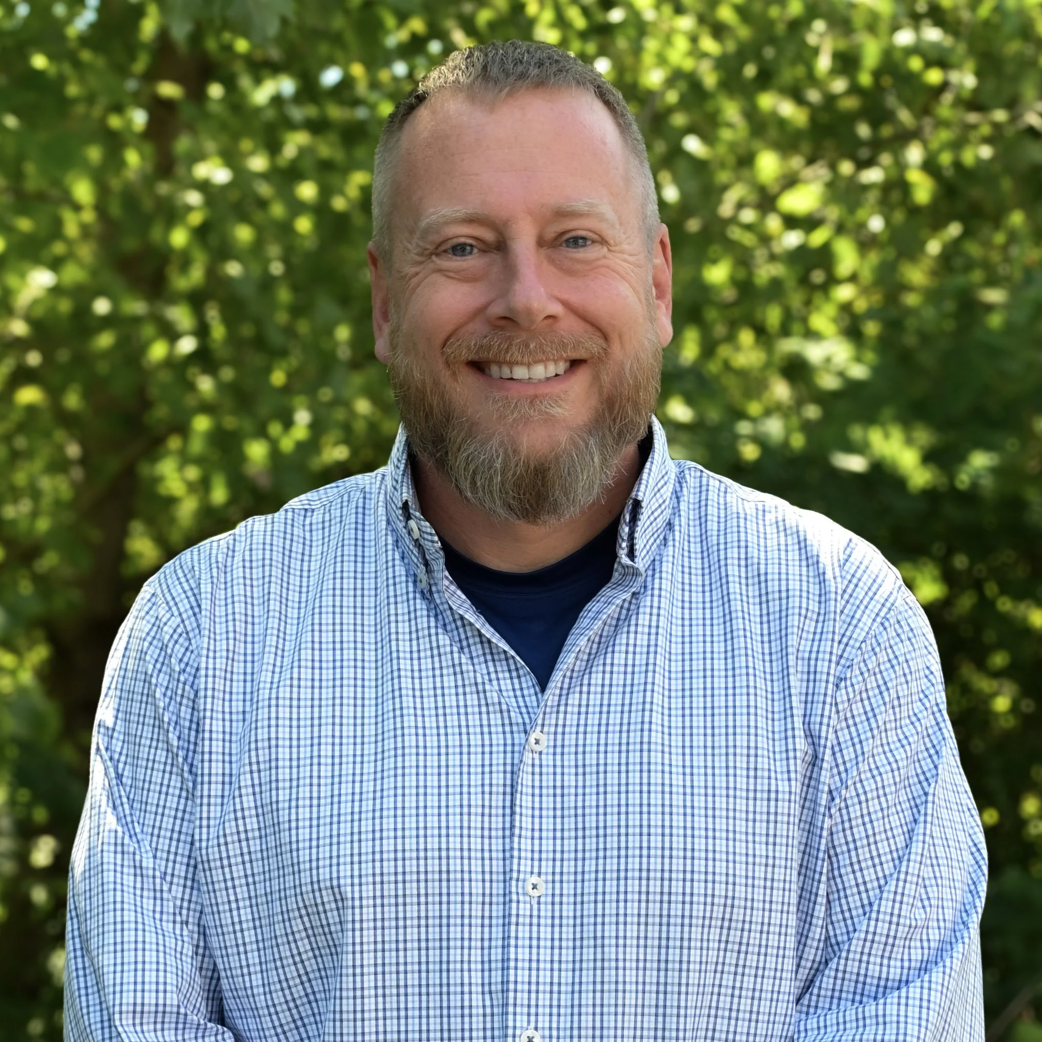 A smiling man with a beard and short hair wearing a checkered shirt, standing outdoors with green trees in the background.