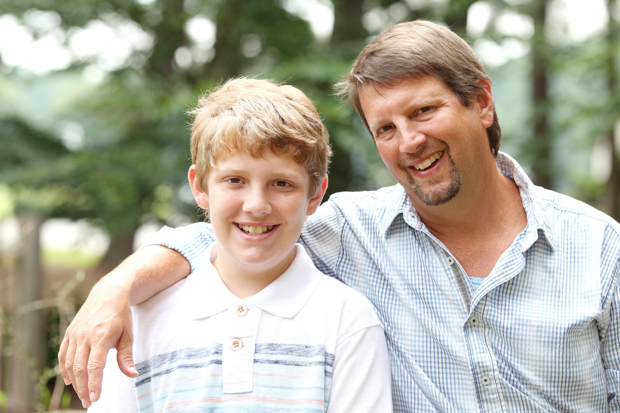 A smiling man with his arm around a young boy, outdoors in a green, wooded area.