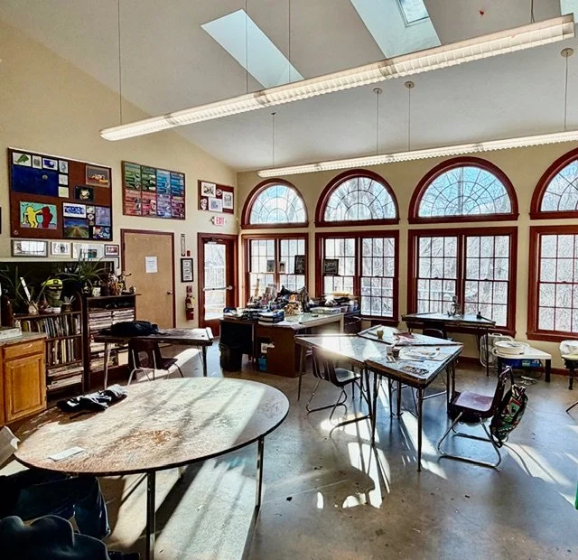 A classroom with large arched windows, desks, chairs, and educational posters on the walls.
