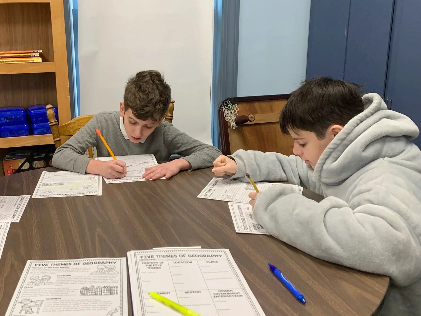 Two boys sitting at a wooden table, working on worksheets about the five themes of geography. One boy is wearing a gray hoodie, and the other is in a gray sweater. There are pens and additional worksheets on the table, and a bookshelf and blue cabinet are visible in the background.