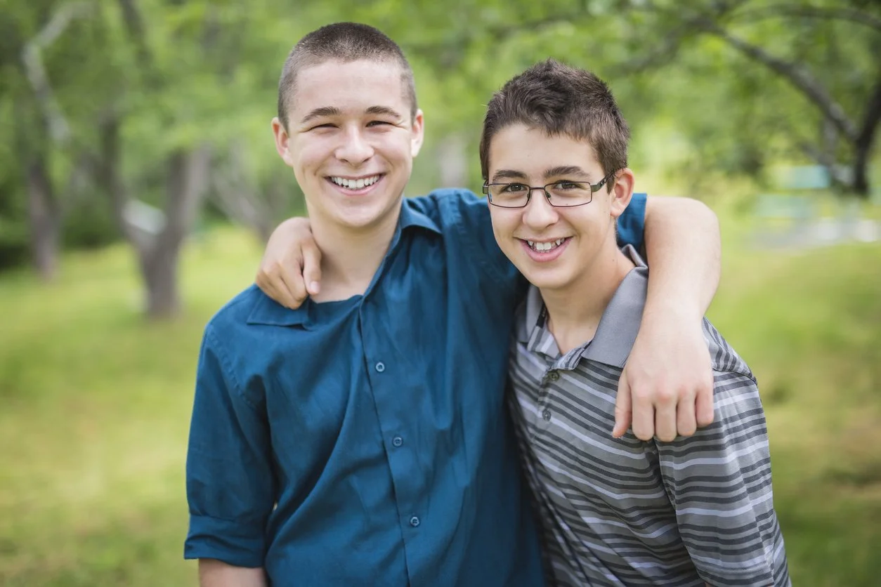 Two teenage boys with light skin smiling, with one having his arm around the other's shoulder, outdoors in a park with green trees in the background.