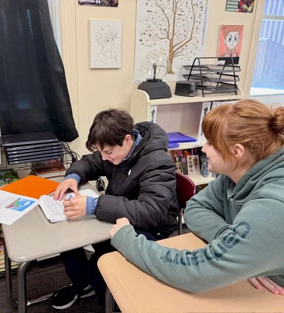 A young boy sitting at a school desk reading a book, with a woman sitting beside him, in a classroom decorated with artwork and a bookshelf.