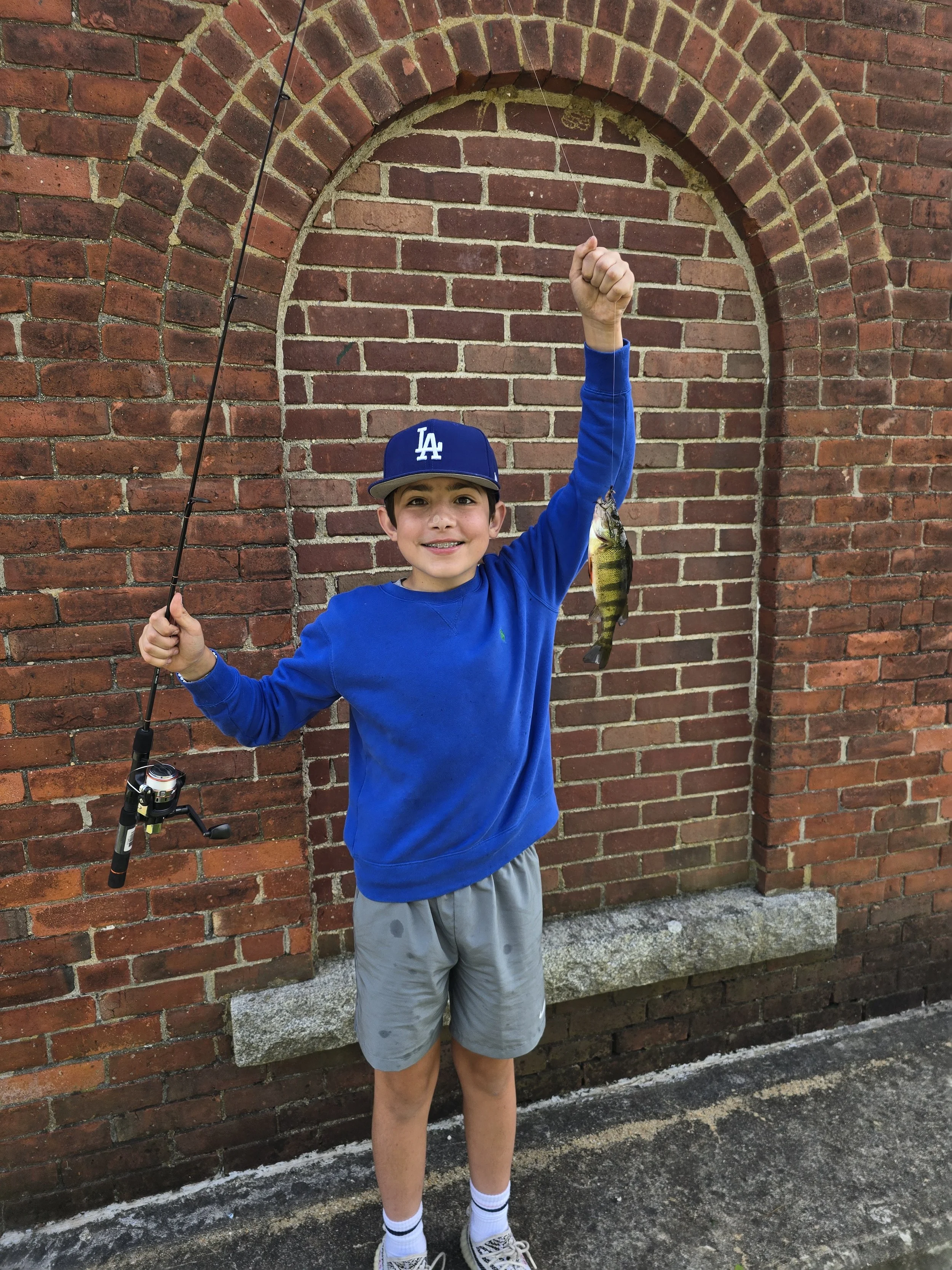 A young boy in a blue sweatshirt, gray shorts, and a blue Los Angeles Dodgers cap is standing in front of a brick wall, holding a fishing rod in his right hand and a small fish in his left hand.