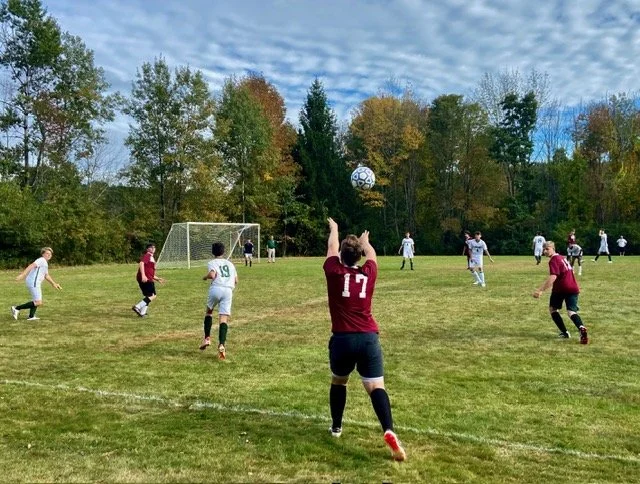 A girls' soccer game on a grassy field with trees in fall colors in the background, players in white and maroon jerseys, some running, with a soccer ball in mid-air.