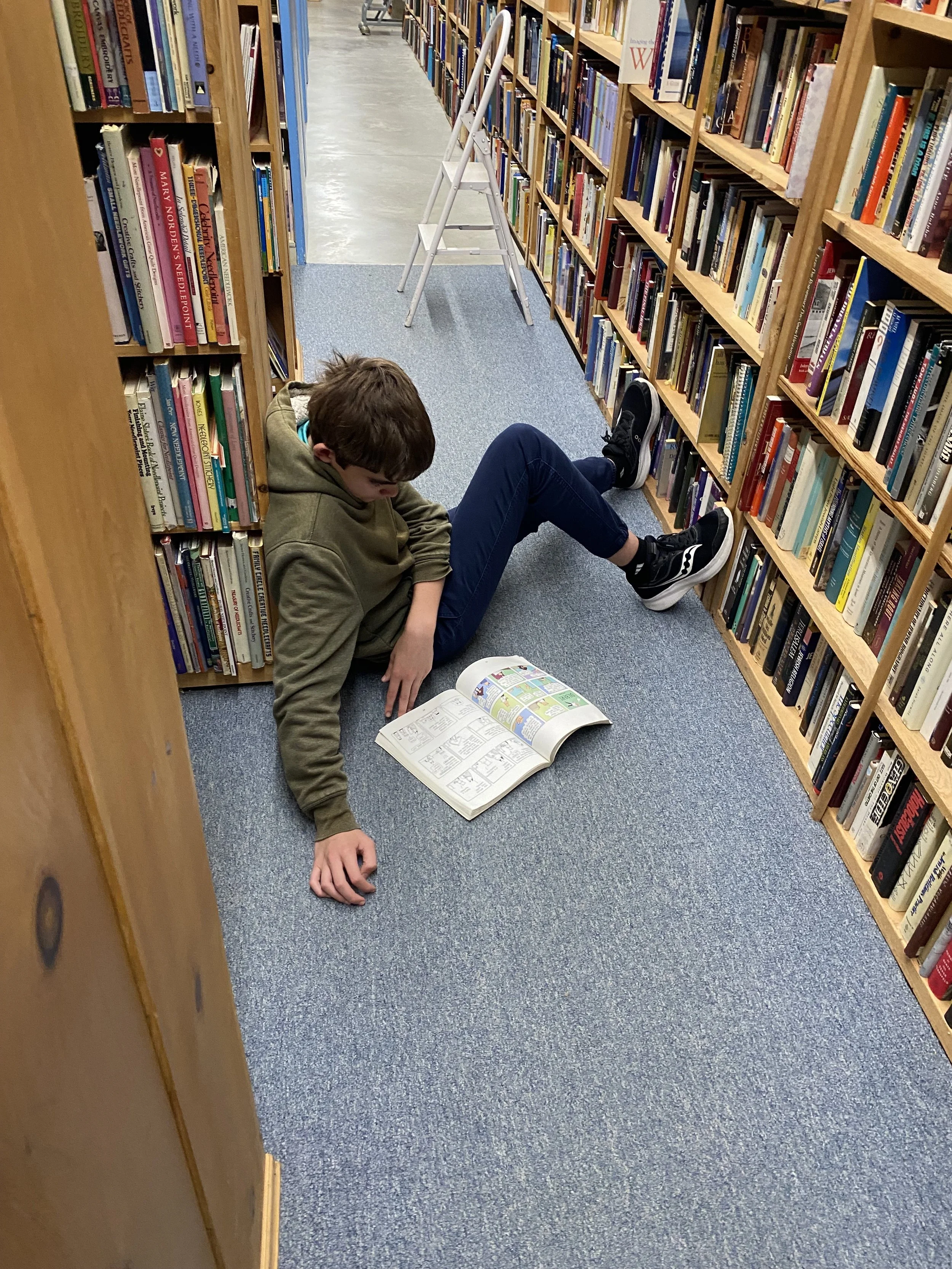 A boy lying on the floor of a library between bookshelves, reading a comic book.
