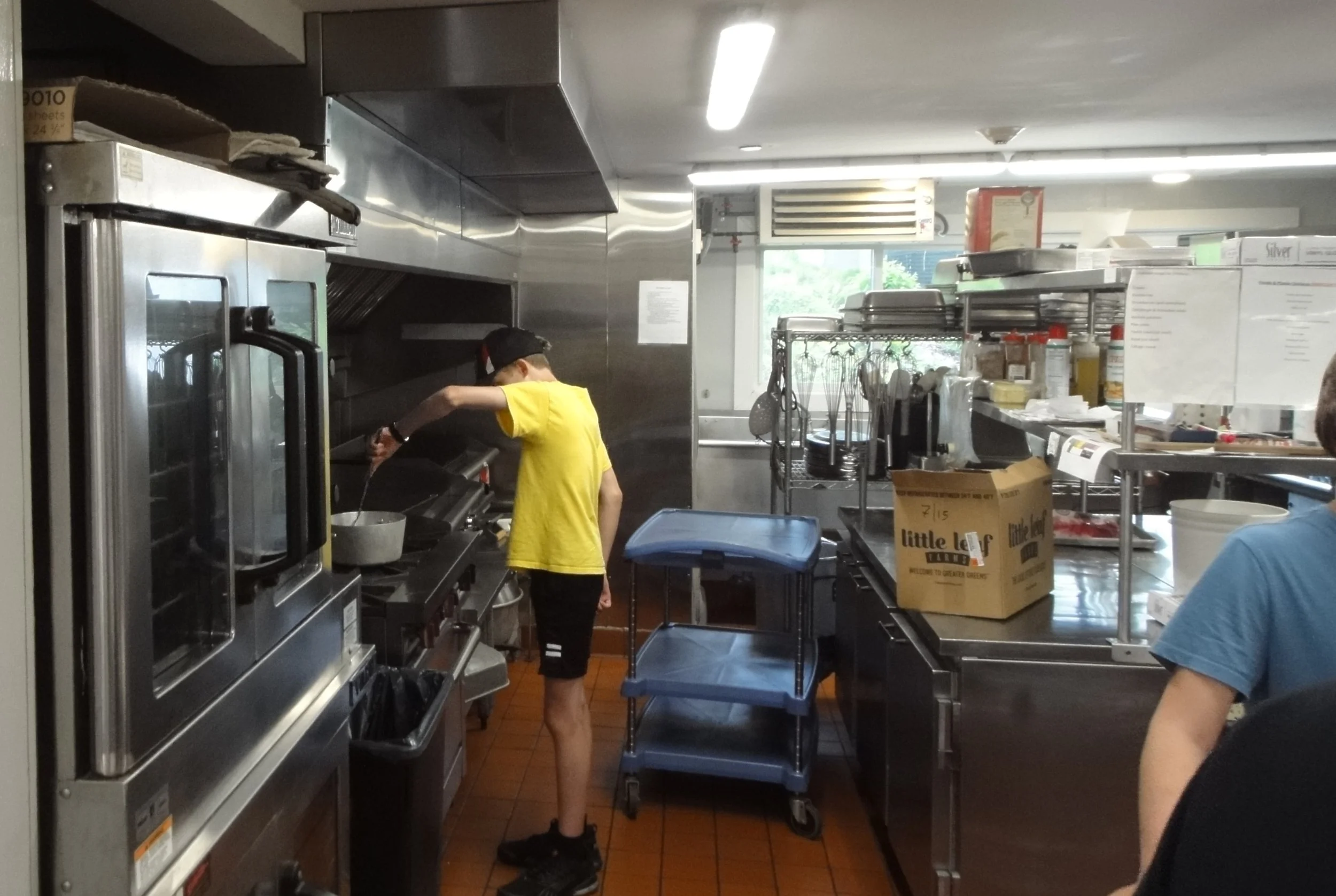 A person in a yellow T-shirt and black shorts cooking on a griddle in a commercial kitchen, with shelves of utensils and supplies in the background.