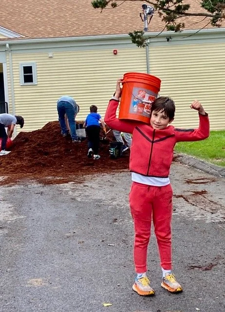 Child in red hoodie and pants holding a large orange bucket, standing on a gravel driveway near a pile of dirt and other children working in the background.