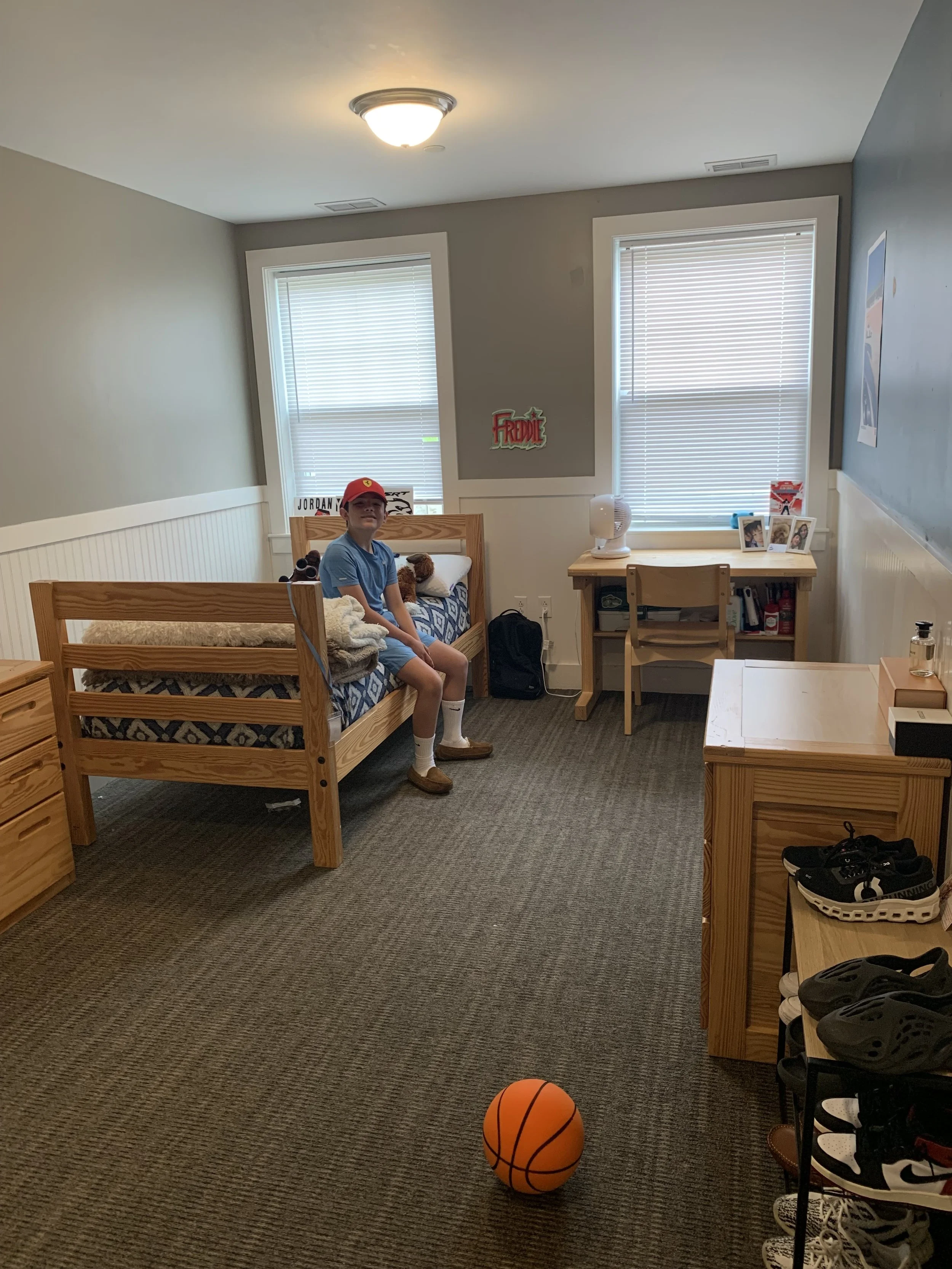 A young boy sitting on a bed in a bedroom, wearing a red cap, blue shirt, shorts, and brown shoes, with toys and a pillow on the bed, and a basketball on the floor.