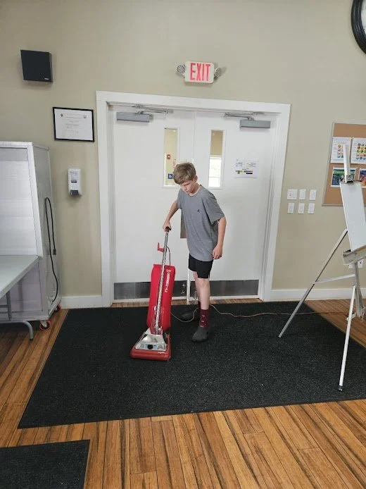 A young boy vacuuming a carpeted area in a room with beige walls, white double doors, and wooden floors.