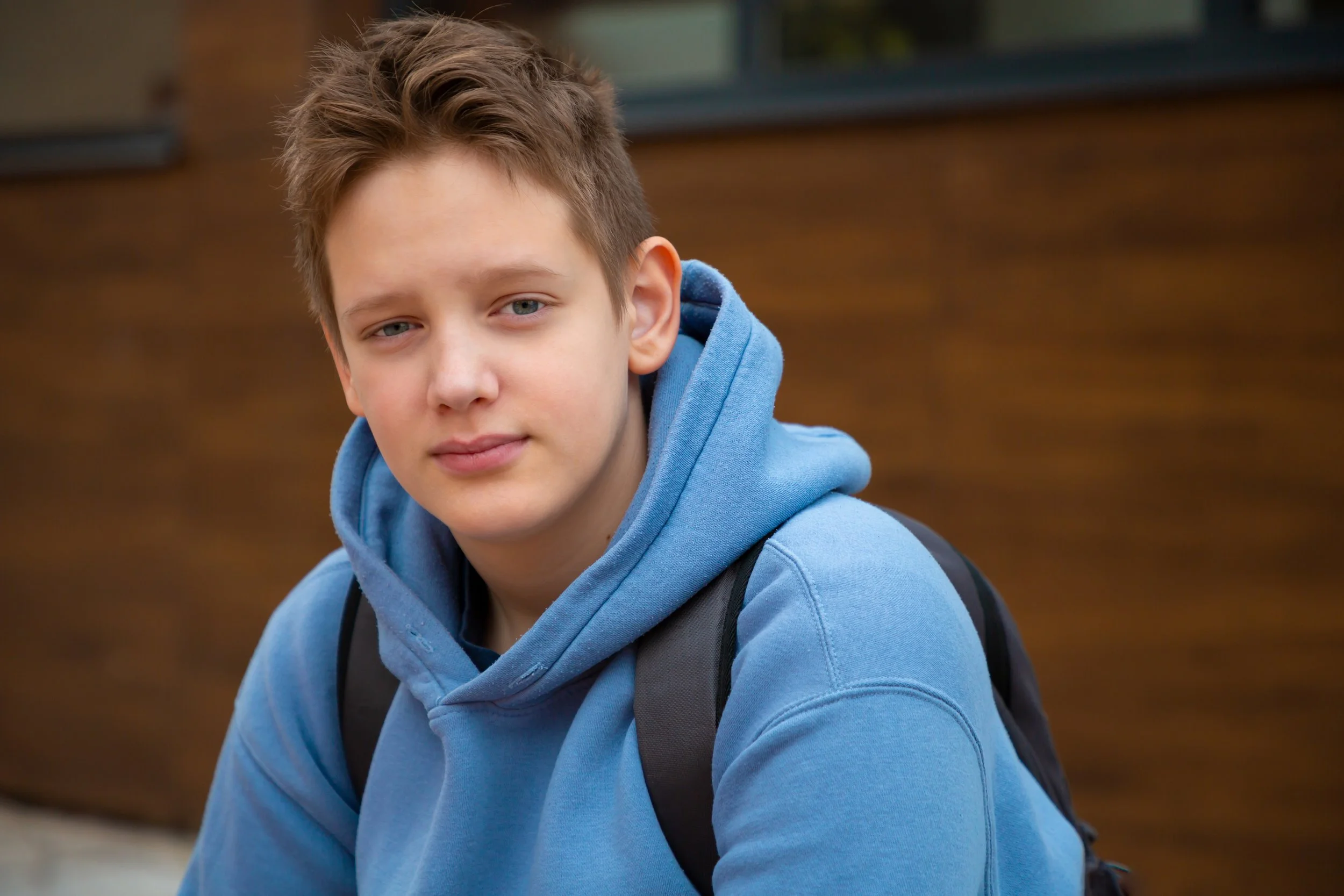 A teenage boy with blue eyes and brown hair wearing a blue hoodie and a backpack outdoors.
