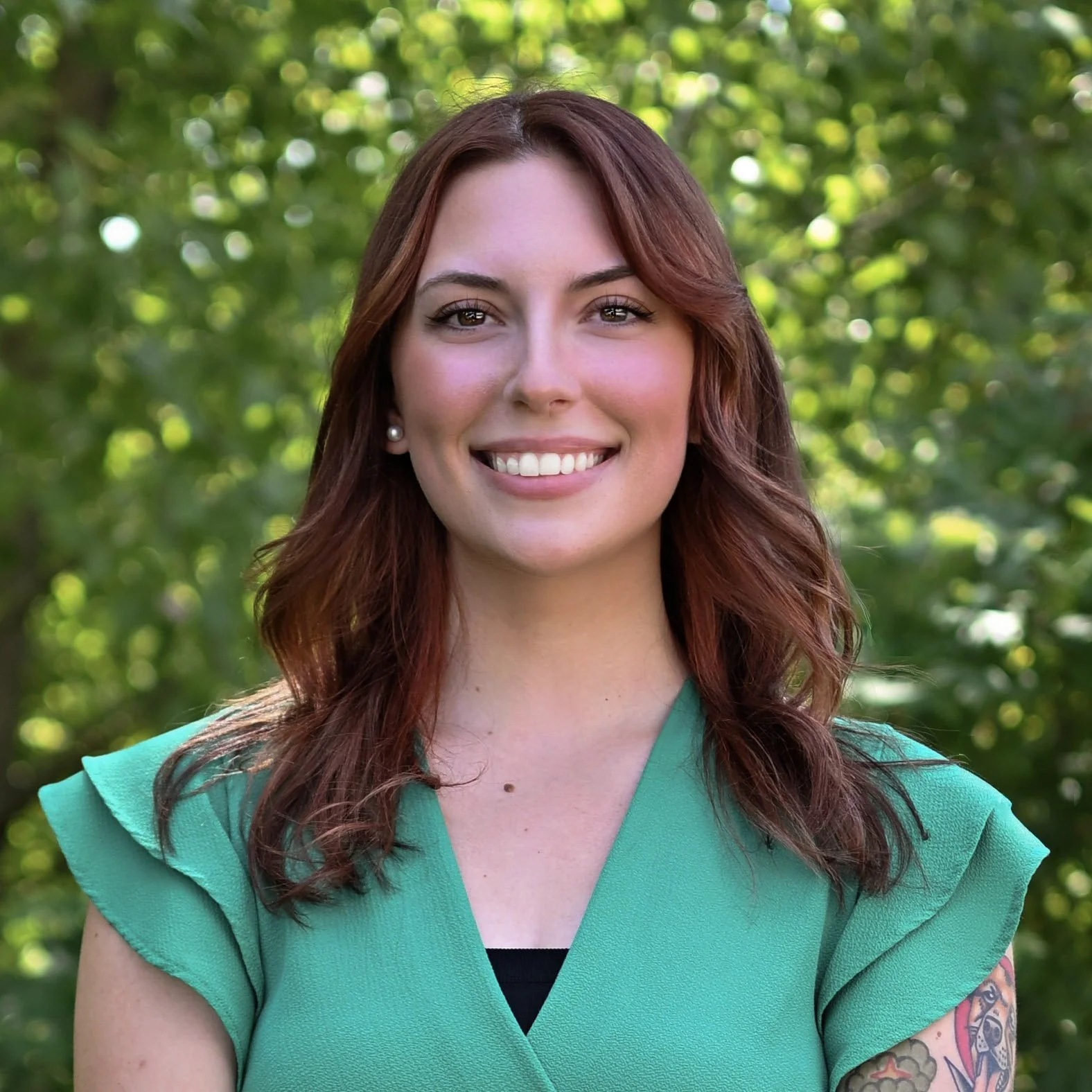 A young woman with shoulder-length wavy auburn hair, smiling with white teeth, wearing pearl earrings and a teal top with ruffled sleeves, standing outdoors with a background of green trees and sunlight.