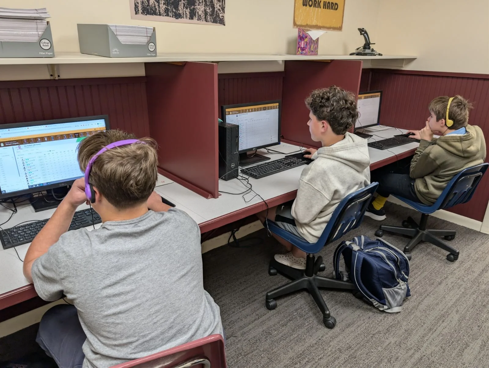 Three boys sitting at computers in a classroom, focusing on their screens; two of them wearing headphones and one with a backpack on the floor beside him.