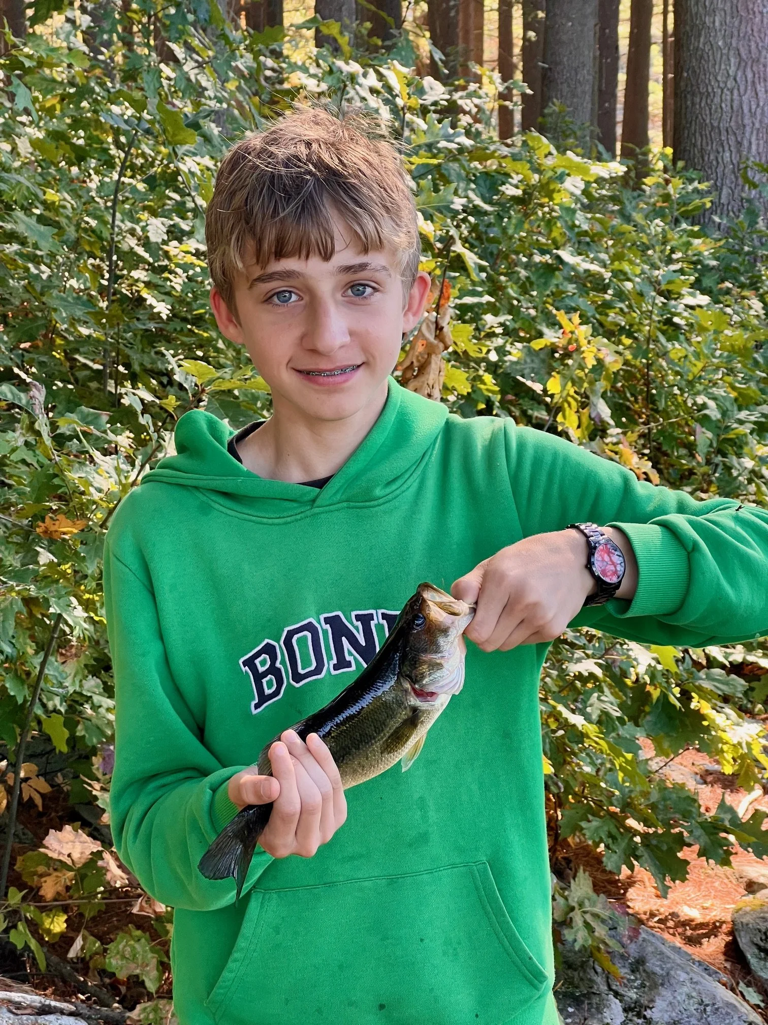A boy with blue eyes and brown hair holding a fish in a forested area.