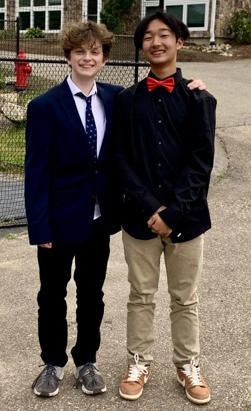 Two young men standing outdoors near a chain-link fence, smiling at the camera. One is wearing a navy blazer, white dress shirt, and dark pants, while the other is dressed in a black shirt, red bow tie, and khaki pants.