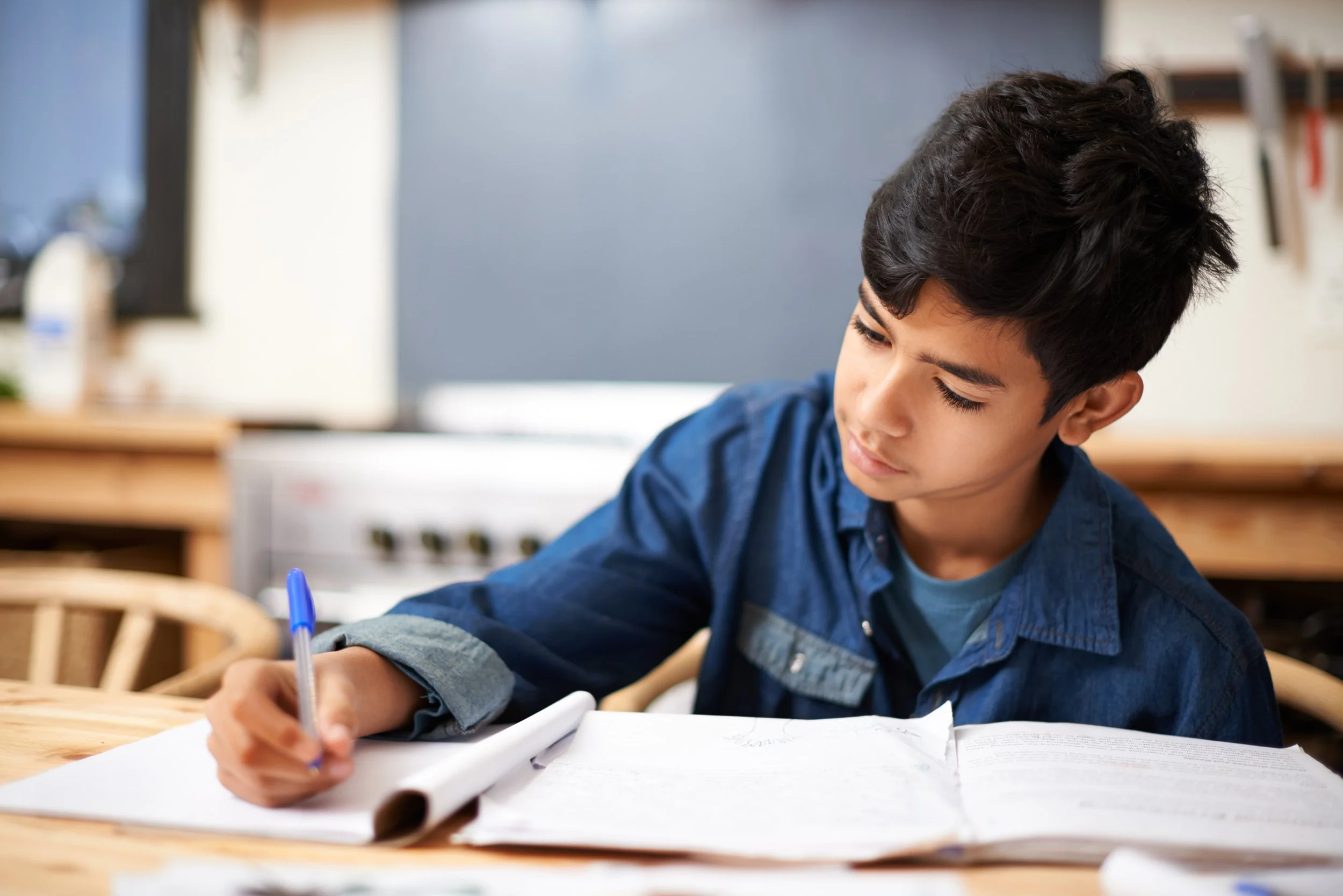 Young boy with dark hair studying at a wooden table with open notebooks and a pen.