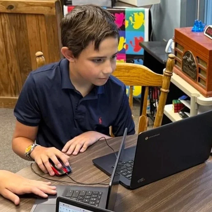 A young boy with brown hair, wearing a navy blue polo shirt, is sitting at a wooden table using a Chromebook laptop and a computer mouse. He appears focused on the screen. The background includes colorful artwork, a vintage radio, and shelves with various items.