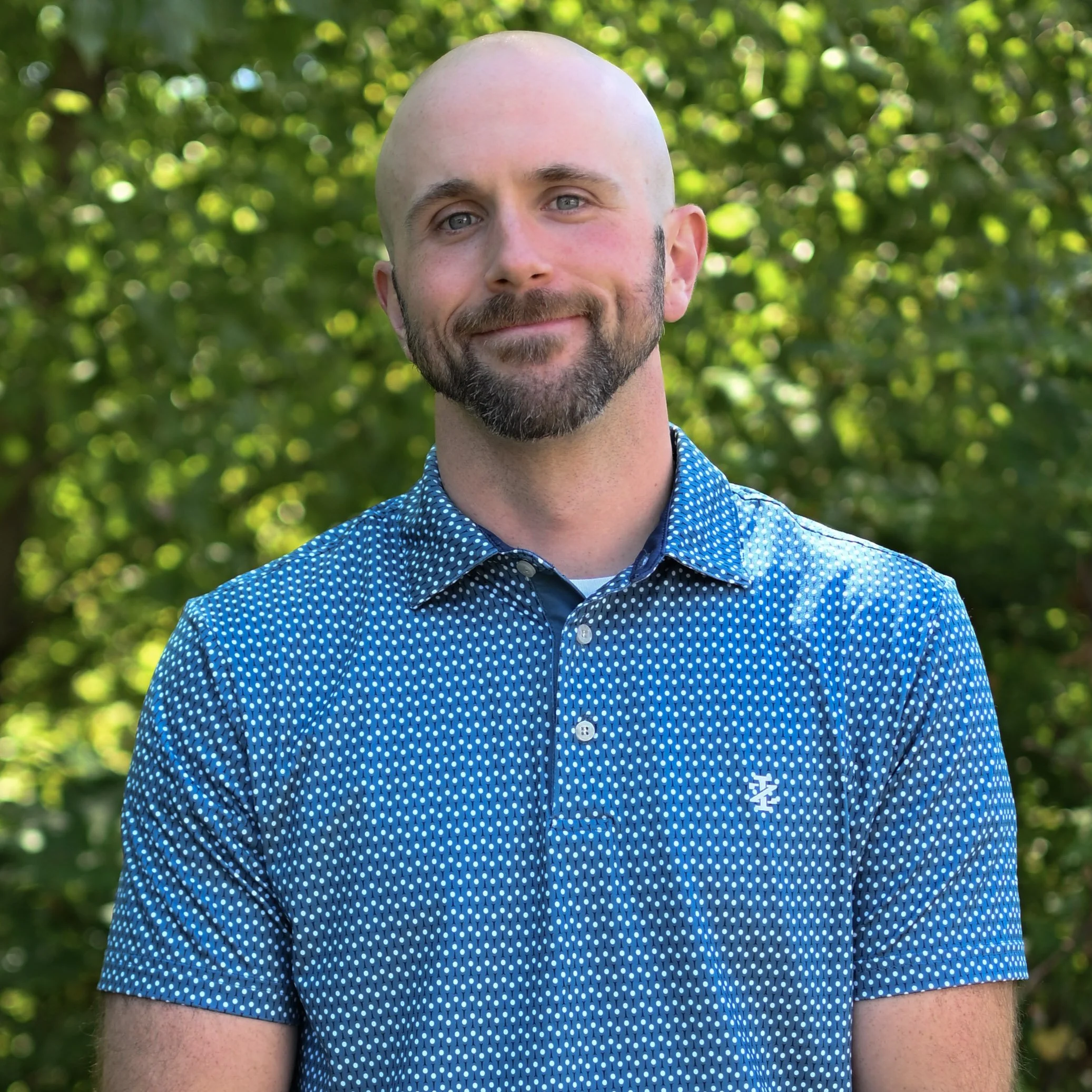 A man with a shaved head and beard smiling outdoors with greenery in the background, wearing a blue patterned short-sleeve shirt.