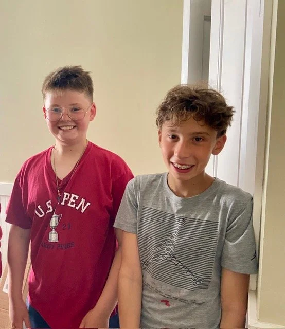 Two boys standing indoors, smiling. One is wearing glasses and a red t-shirt with a chess piece design, the other is in a gray t-shirt. They appear happy and casual.