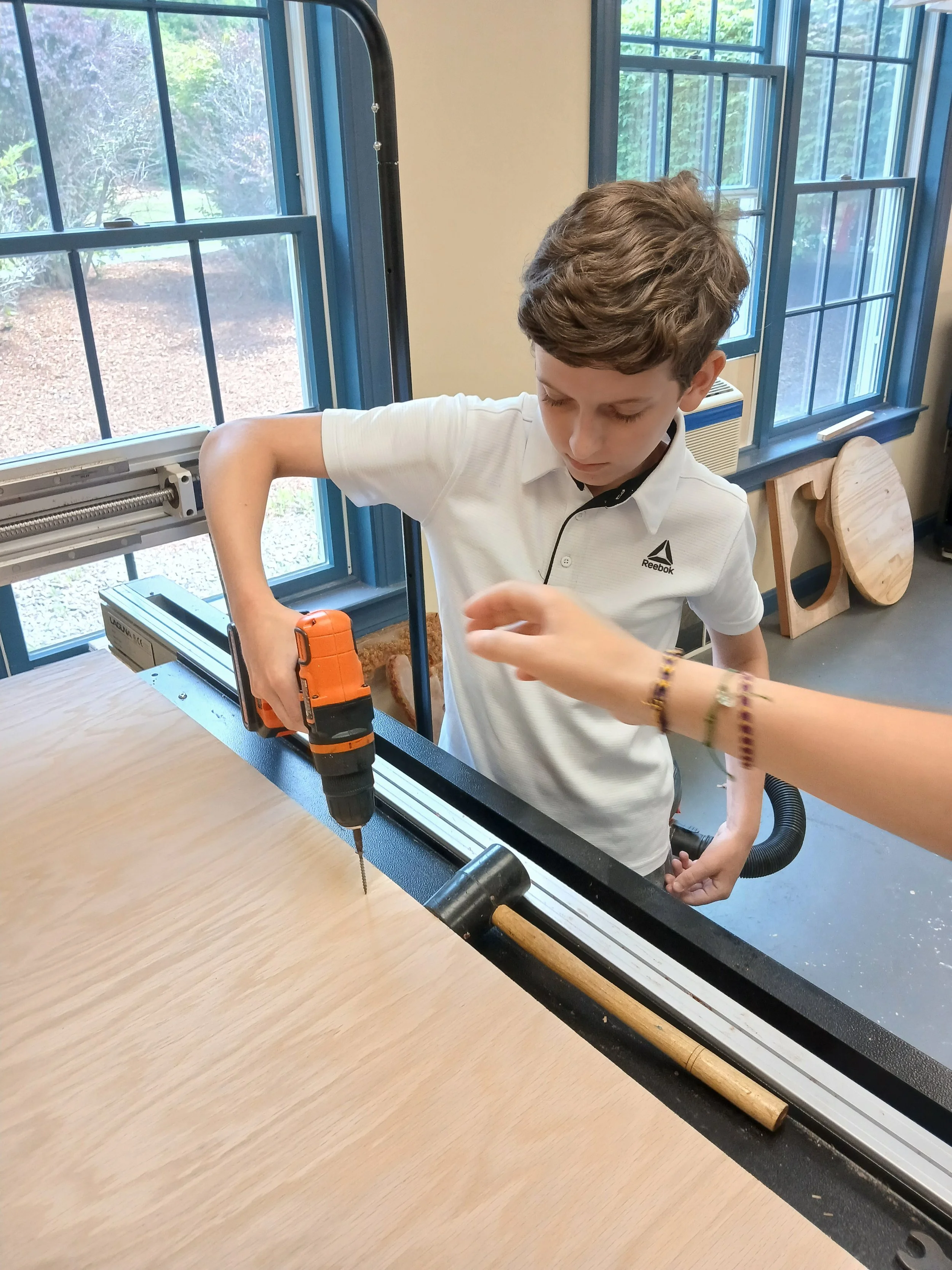 A young boy using a cordless drill to drill into a piece of wood in a workshop with large windows and woodworking tools around.