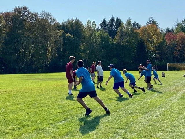 Children playing football on a grassy field with trees in the background.