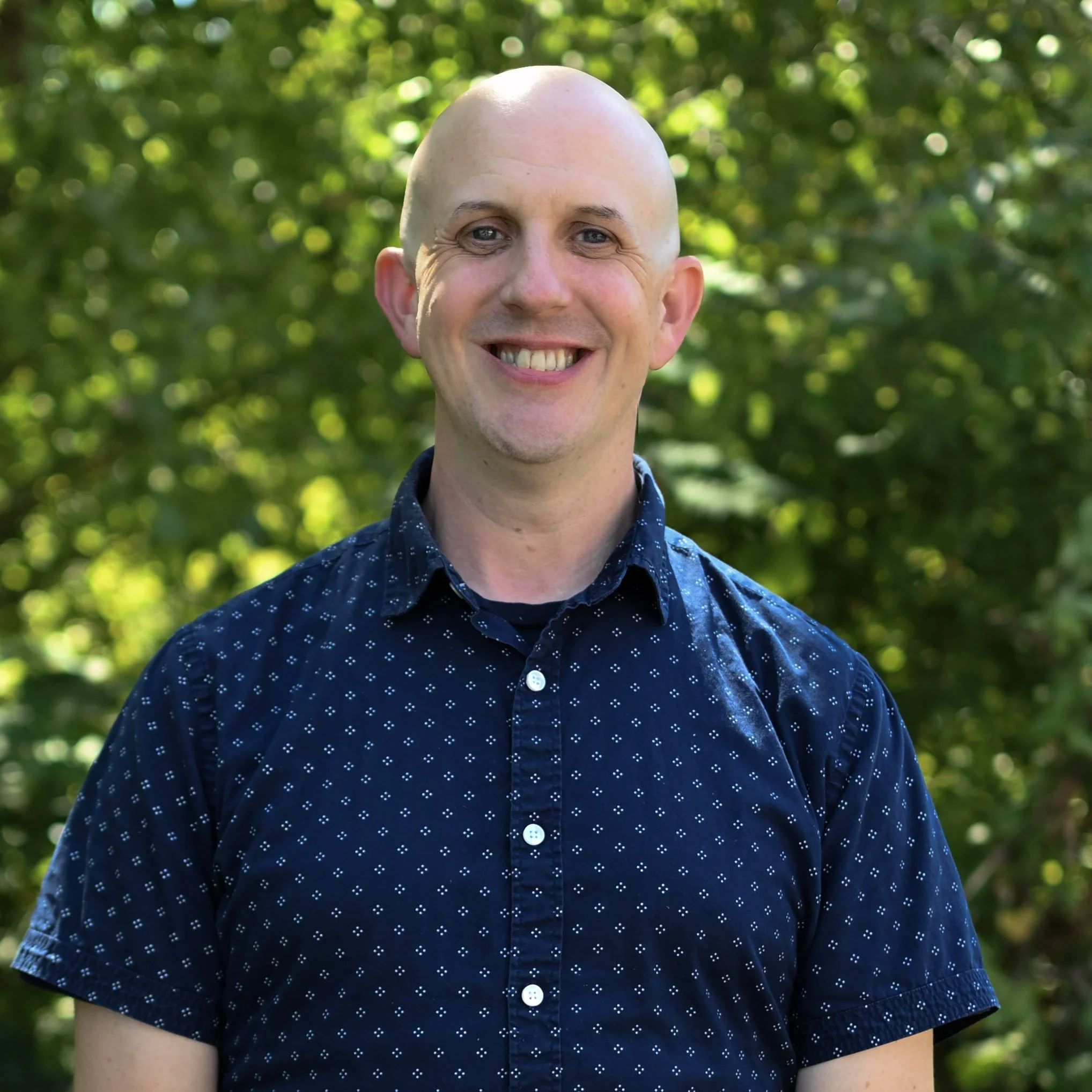 A smiling man with a bald head wearing a navy blue collared shirt with small white patterns, standing outdoors with green foliage in the background.