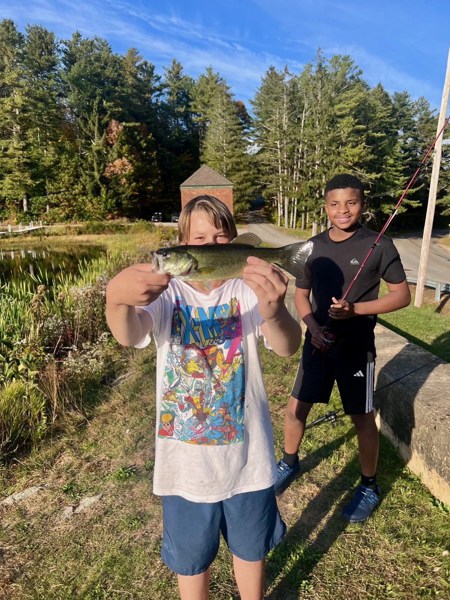 Two boys standing outdoors, one holding a large fish. The boy in the foreground is smiling and displaying the fish, while the boy in the background holds a fishing rod and smiles. There are trees, a small pond or lake, and a building in the background.