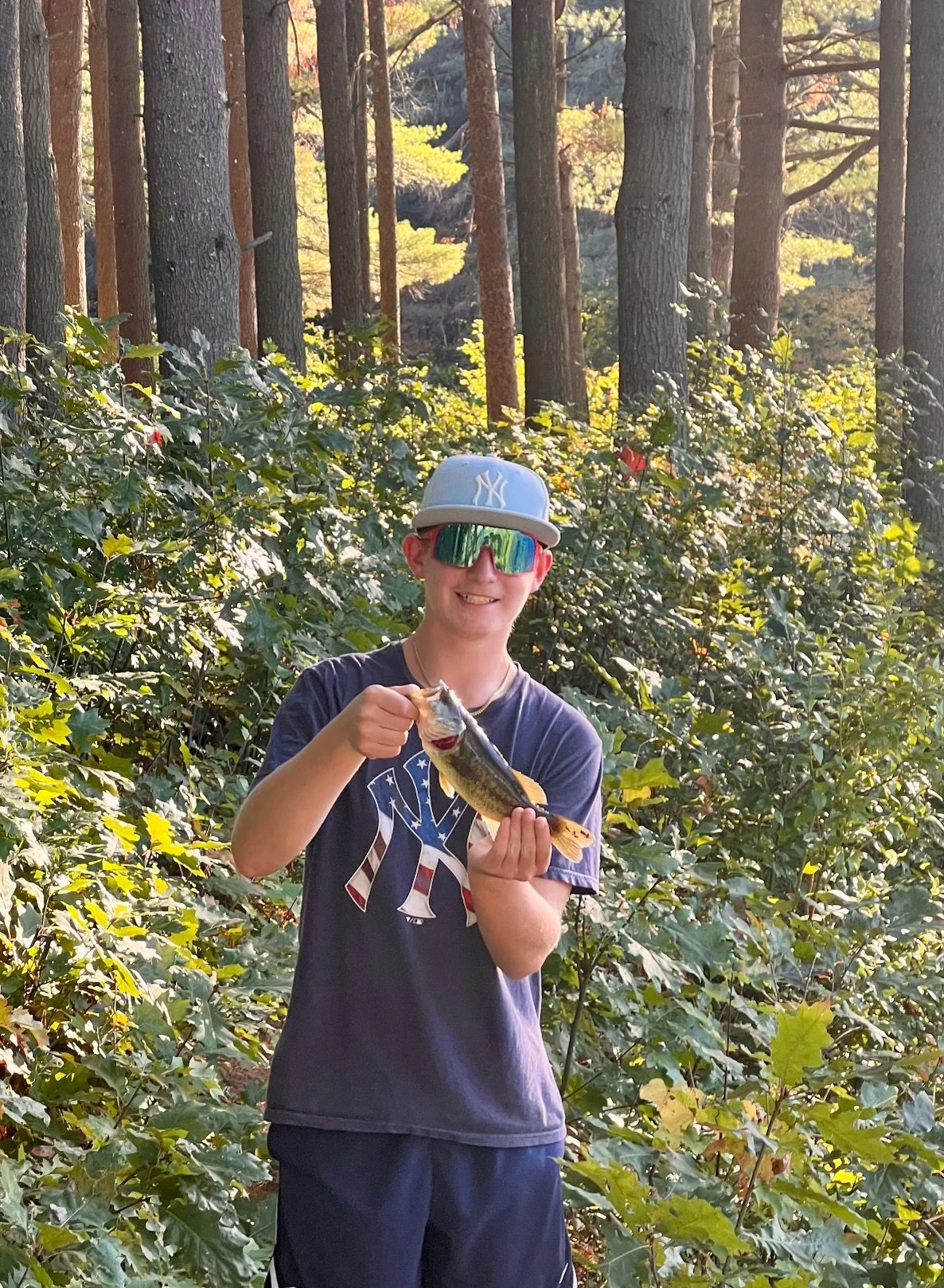 A boy in a New York Yankees baseball cap, sunglasses, and a T-shirt is holding a fish in a forested area with tall trees and green foliage.