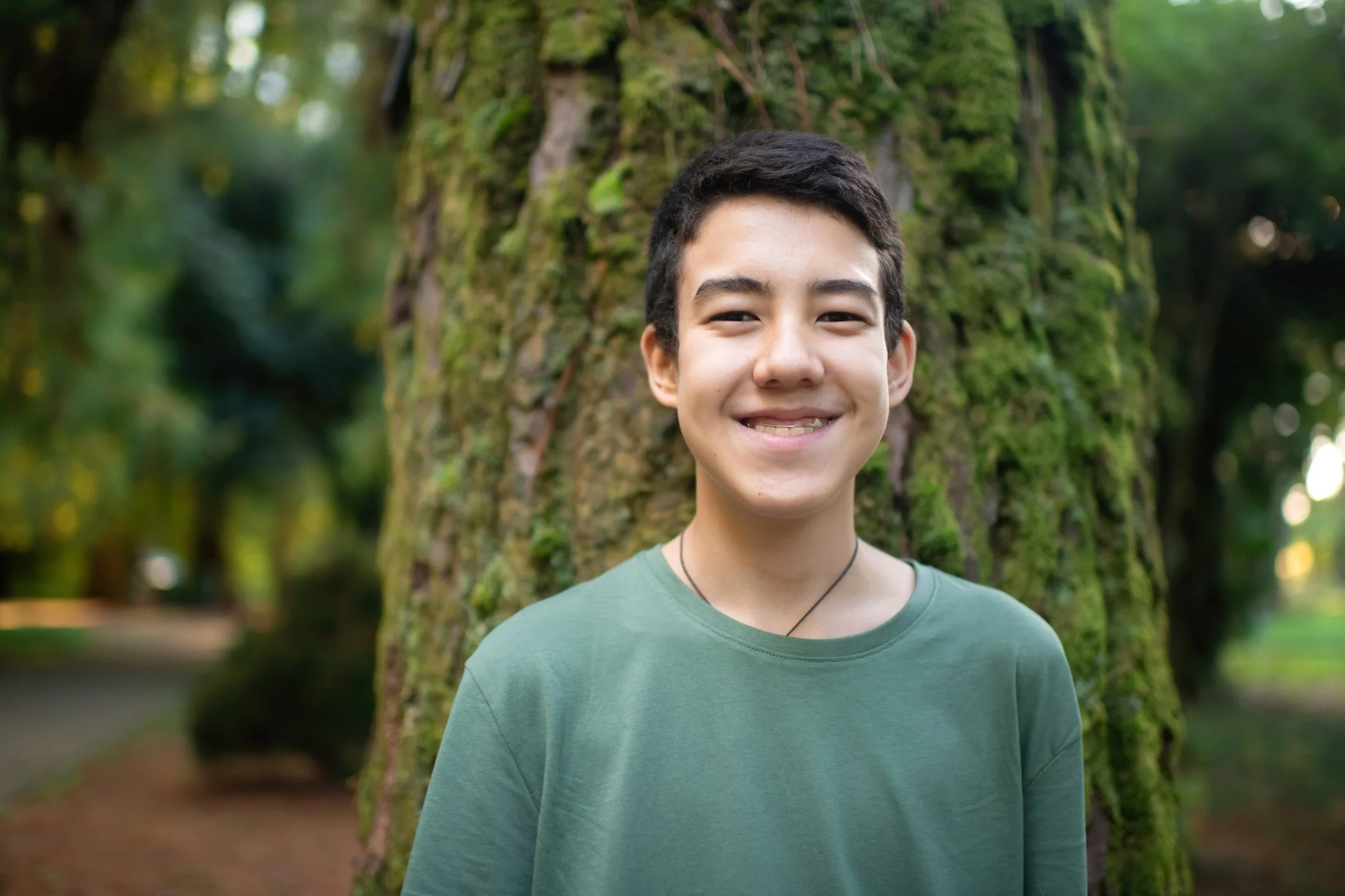 A smiling young person standing near a moss-covered tree in a forest.
