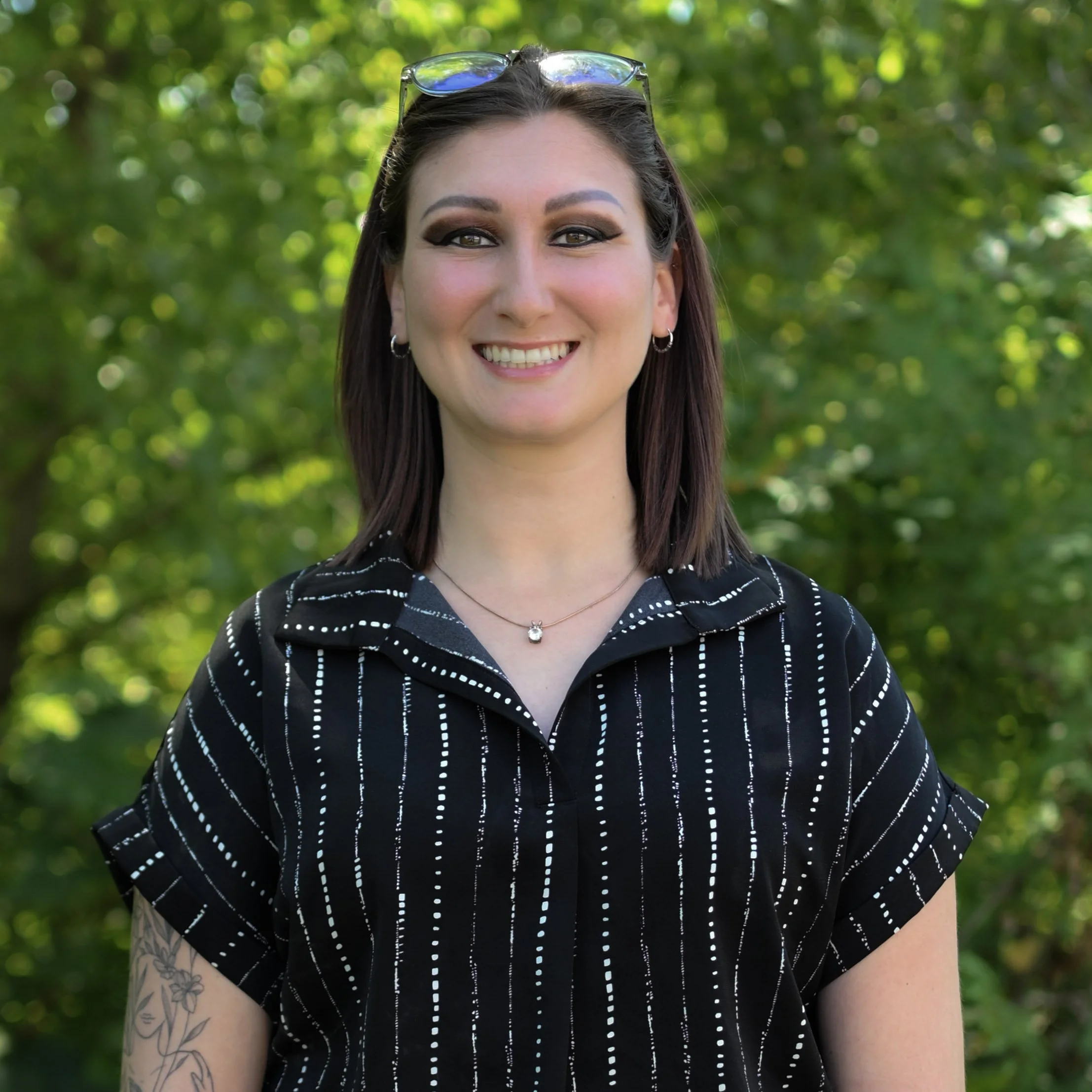 A smiling woman standing outdoors with green trees in the background, wearing sunglasses on her head, a black shirt with white vertical dotted lines, and a silver necklace with a small pendant.