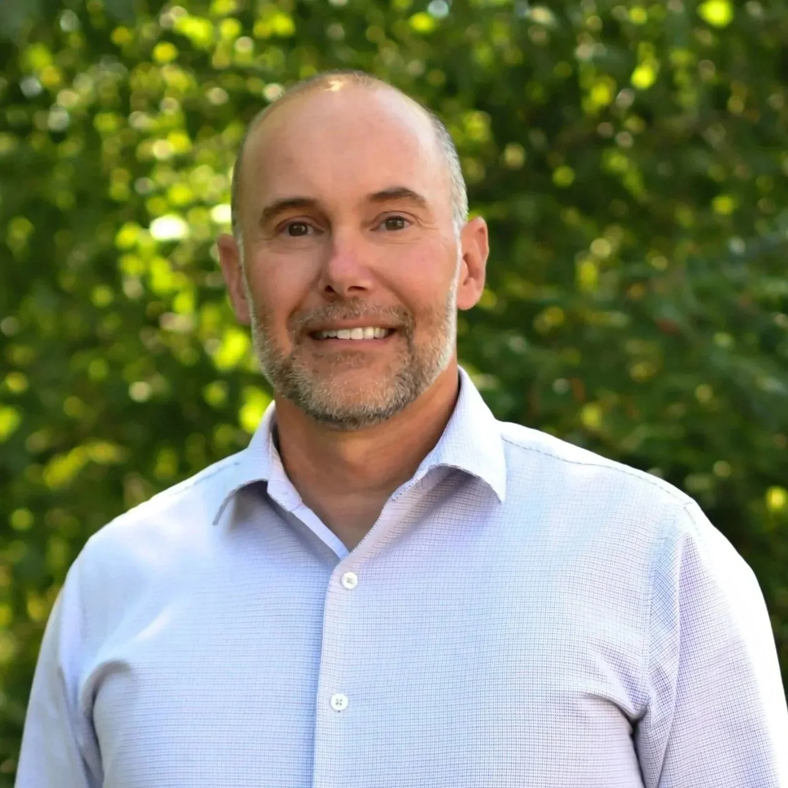 A middle-aged man with a bald head and gray beard, wearing a white dress shirt, standing outdoors with a lush green background.