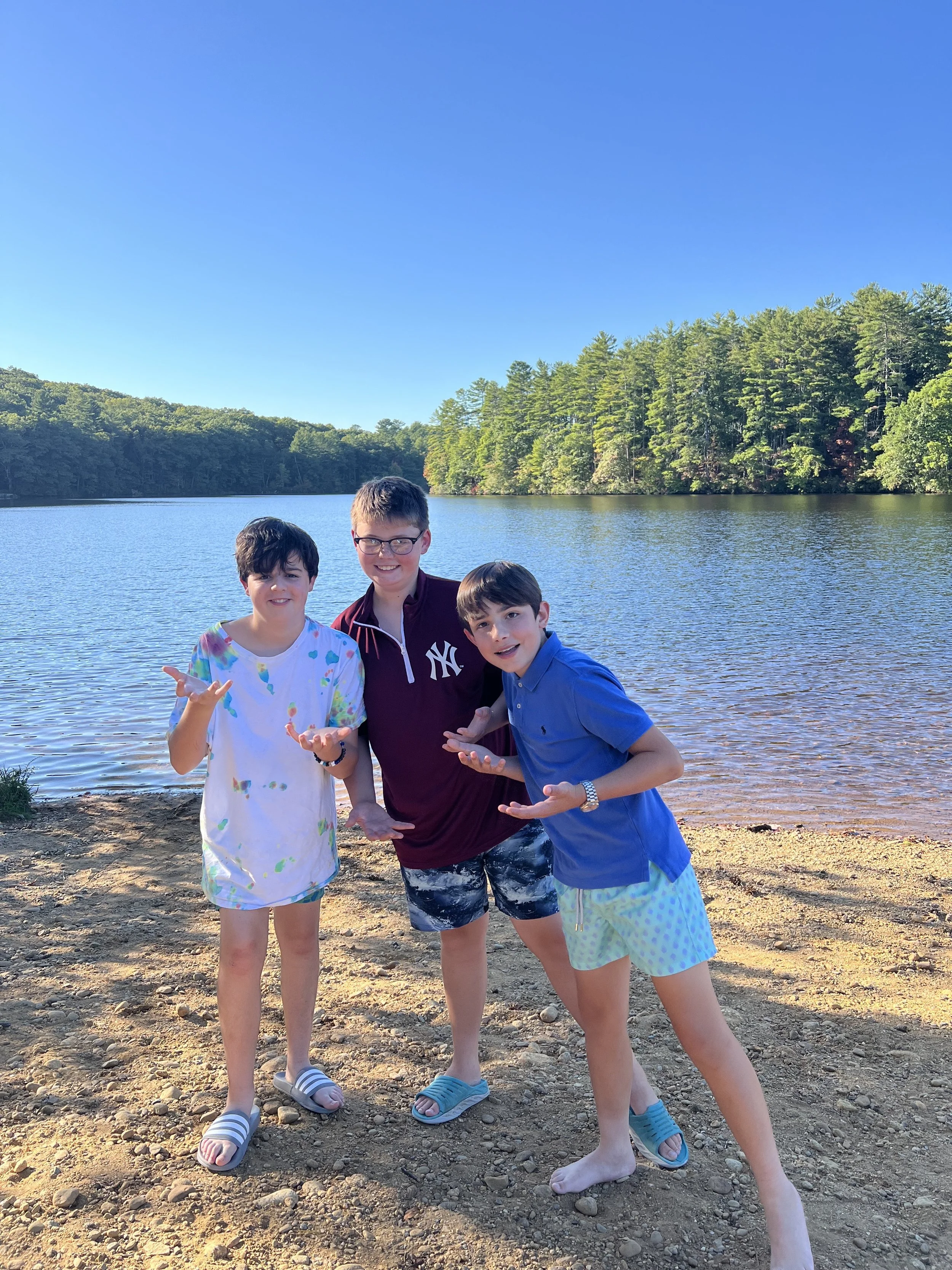 Three children standing on a sandy beach by a lake, with trees and blue sky in the background.