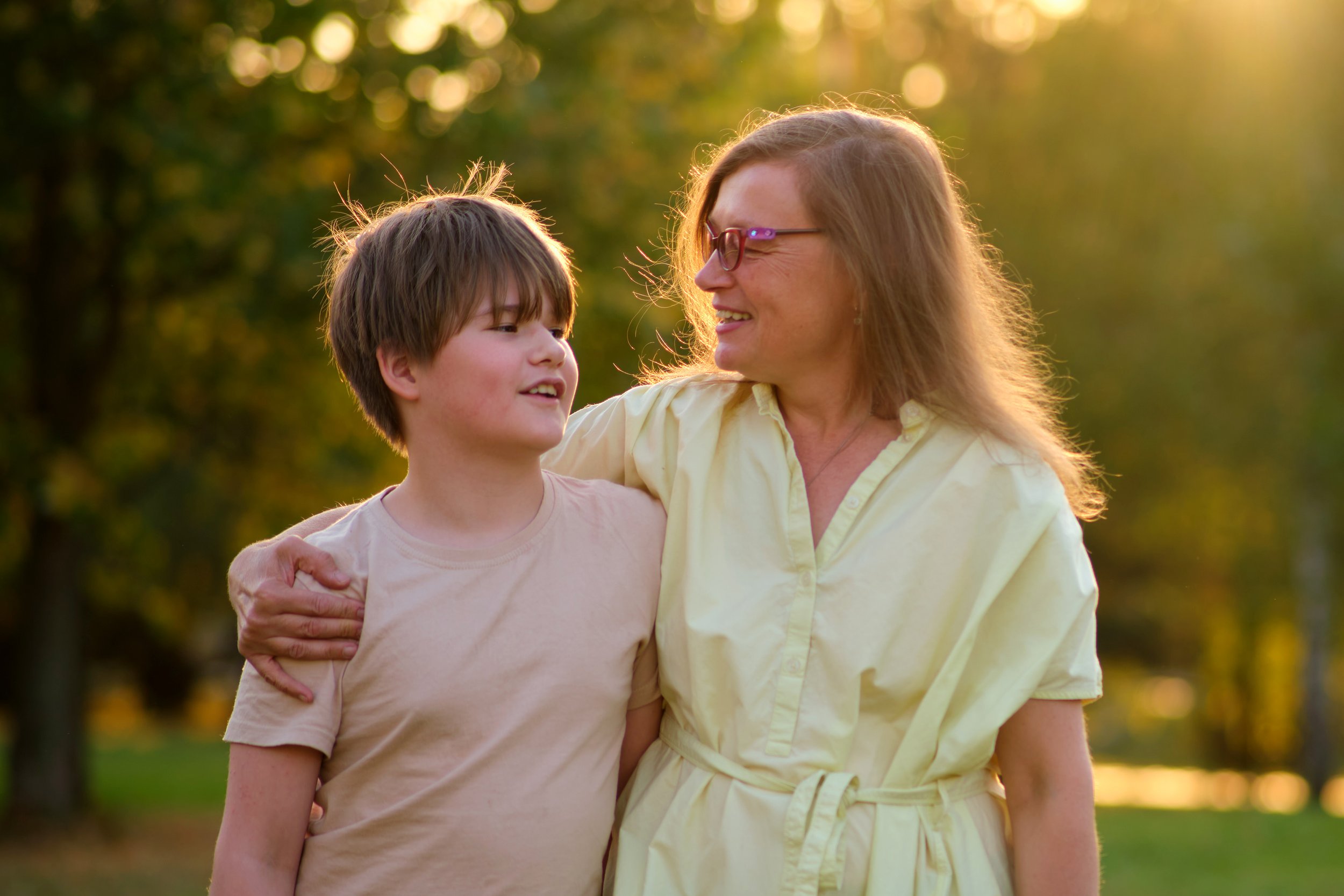 A woman and a young boy smiling and walking together outdoors in a park during sunset, with trees in the background.