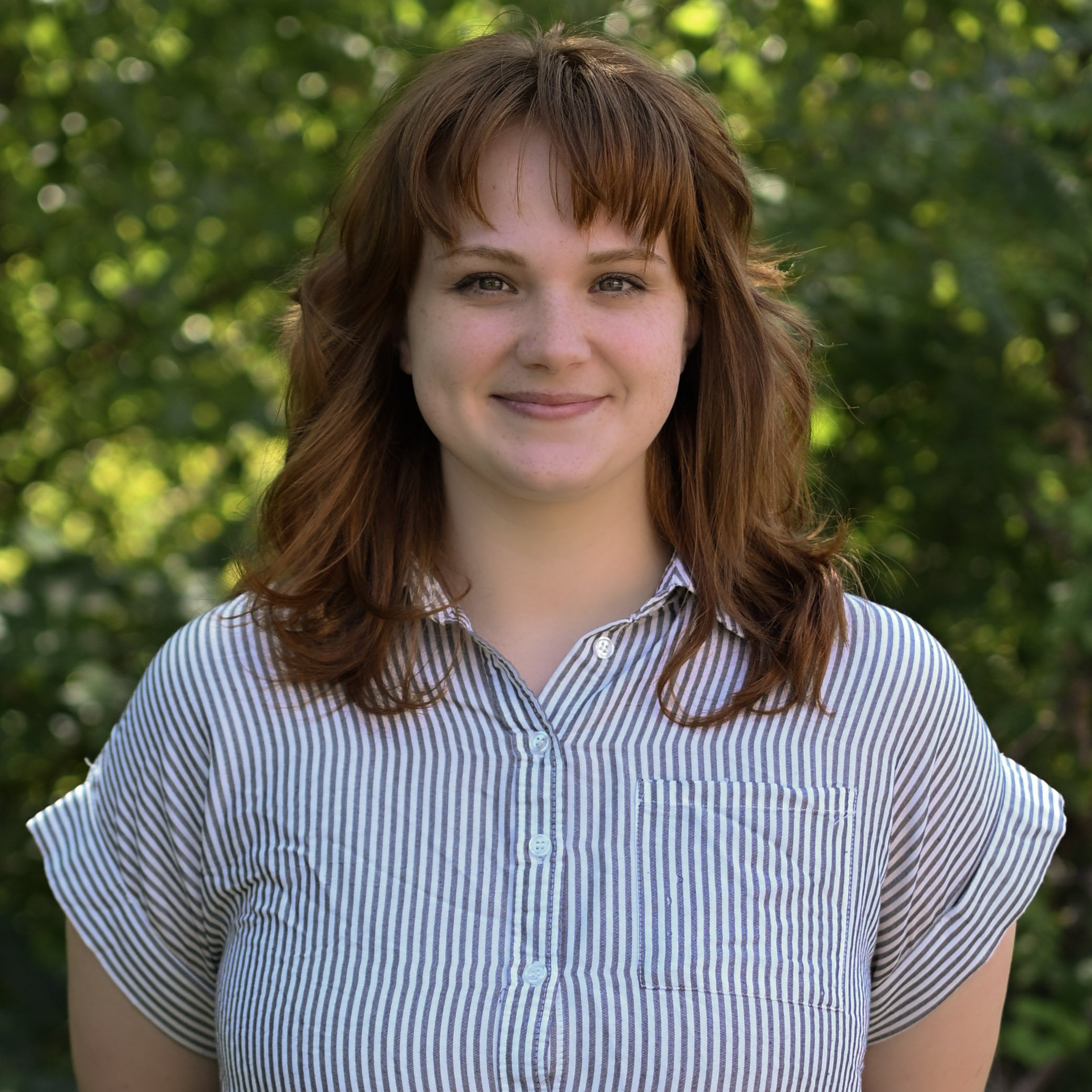 A young woman with red hair and freckles wearing a striped shirt, standing outdoors with green foliage in the background.