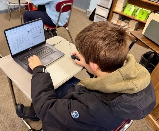 A young boy with brown hair in a black jacket, sitting at a desk in a classroom, working on a laptop and using a computer mouse.