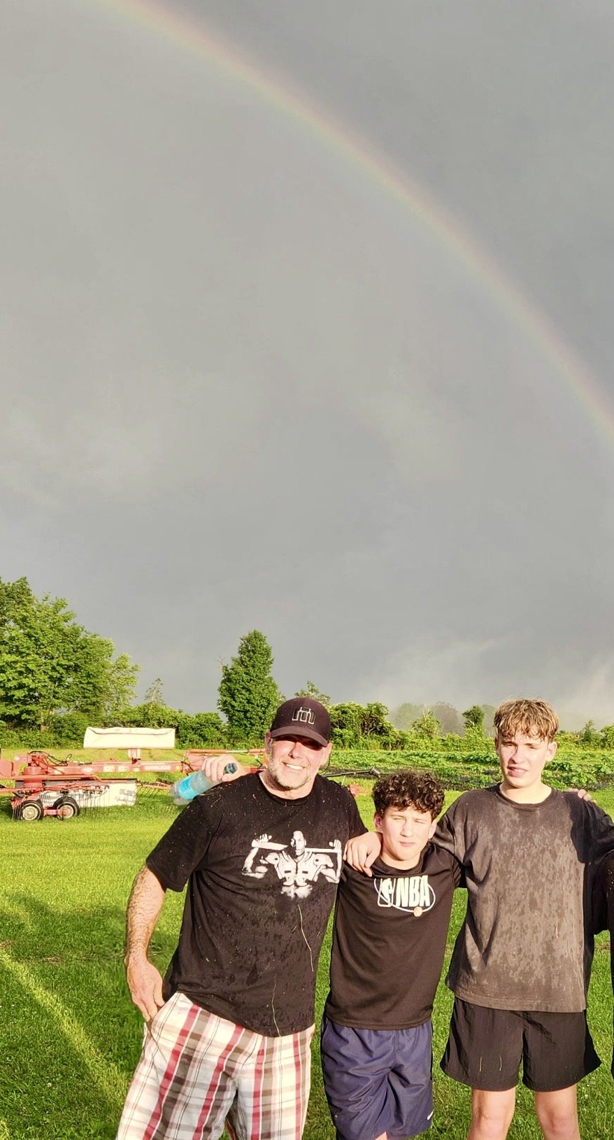 Three people standing outdoors in a field with a rainbow in the cloudy sky. They are smiling and have their arms around each other, with farm equipment and green trees in the background.