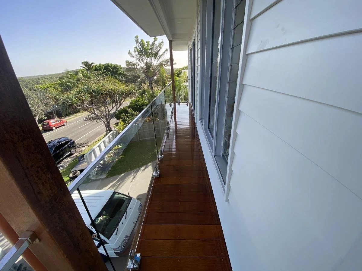 View from a balcony with wooden flooring, glass railing, and sliding glass doors, overlooking a street with cars, trees, and greenery in the background.