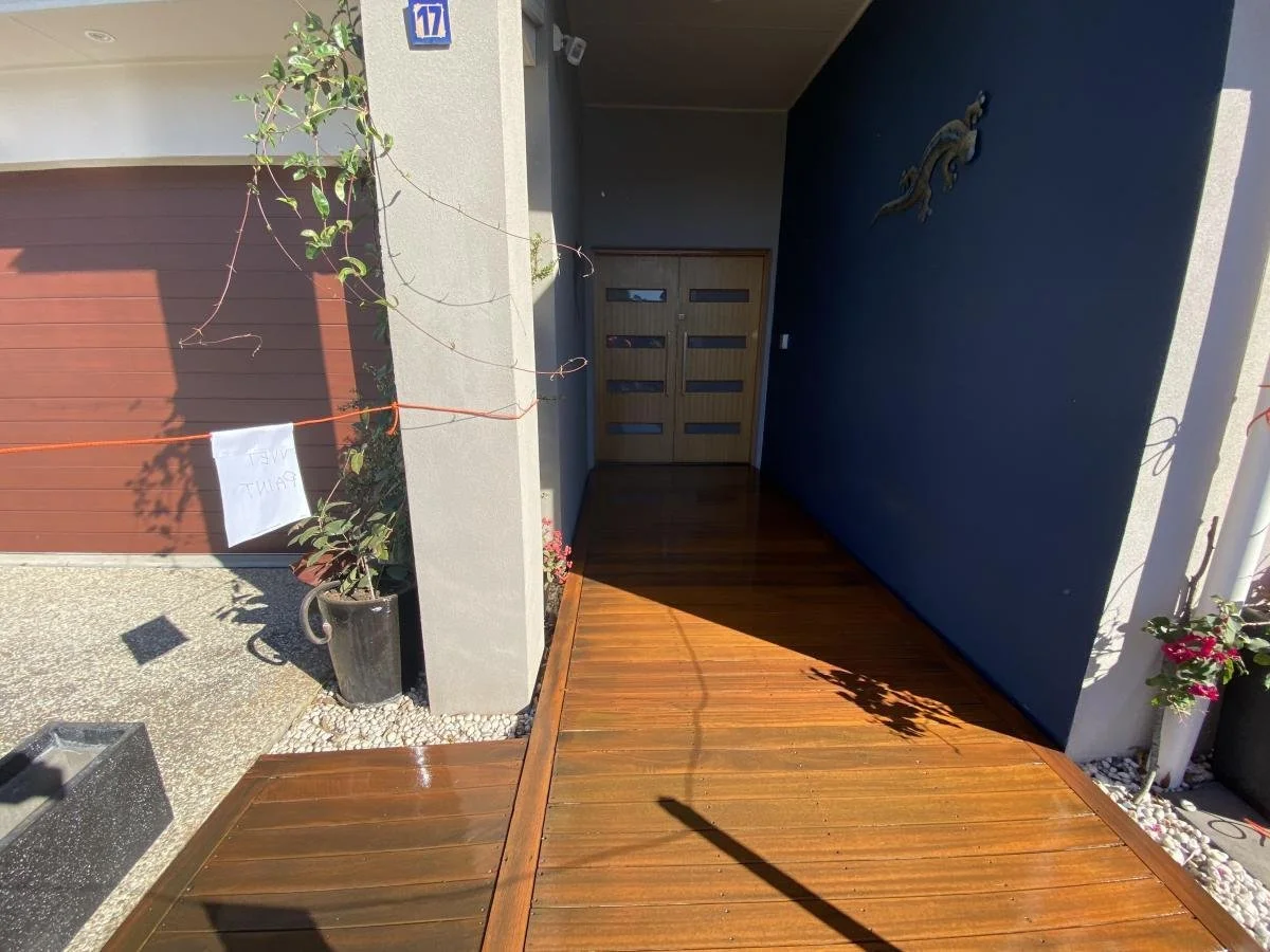 Entrance of a modern house with a wooden deck, a dark blue wall, a metal lizard sculpture, potted plants, and a set of double wooden doors. House number 17 is visible.