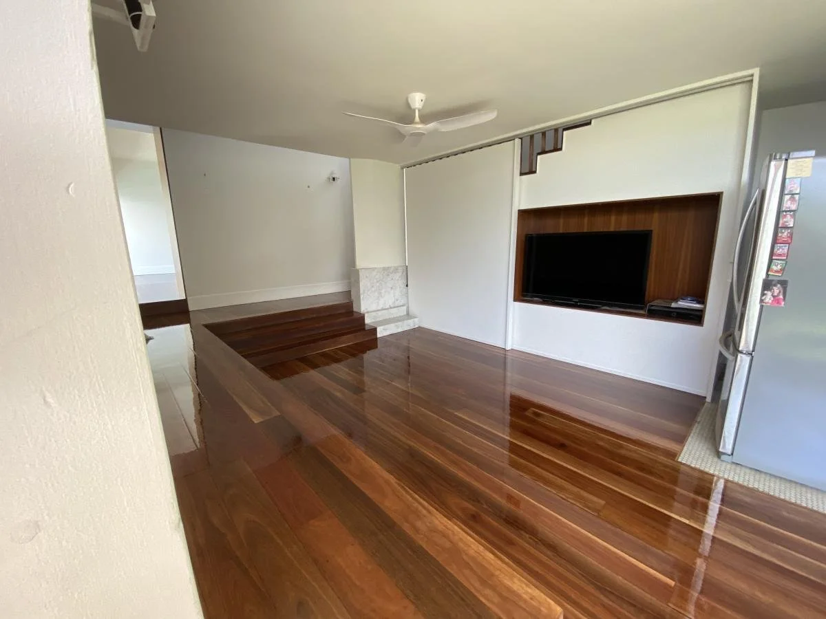 Living room with polished wooden floors, a ceiling fan, a television mounted on a wall with a wooden frame, and a stainless steel refrigerator beside the wall.