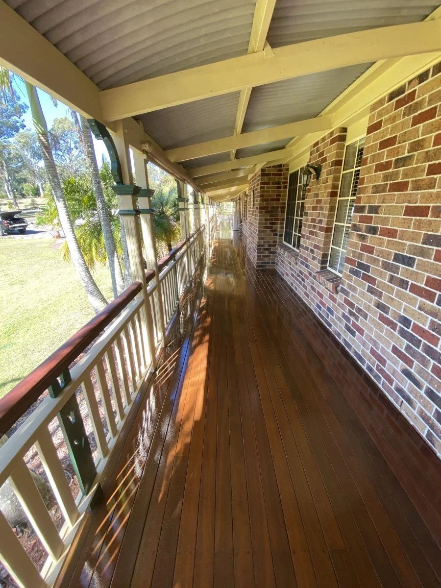 Long wooden porch with beige railing and brick wall, shaded by a roof with exposed beams, outside view includes trees and parked cars.