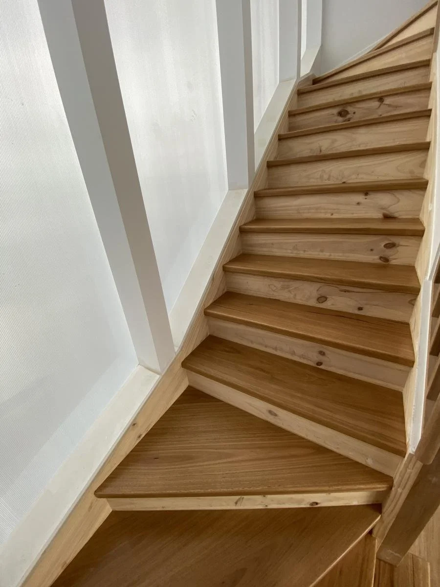 Wooden staircase with a sharp turn, with the top of the staircase leading upward and the bottom turning to the side, set against white walls.